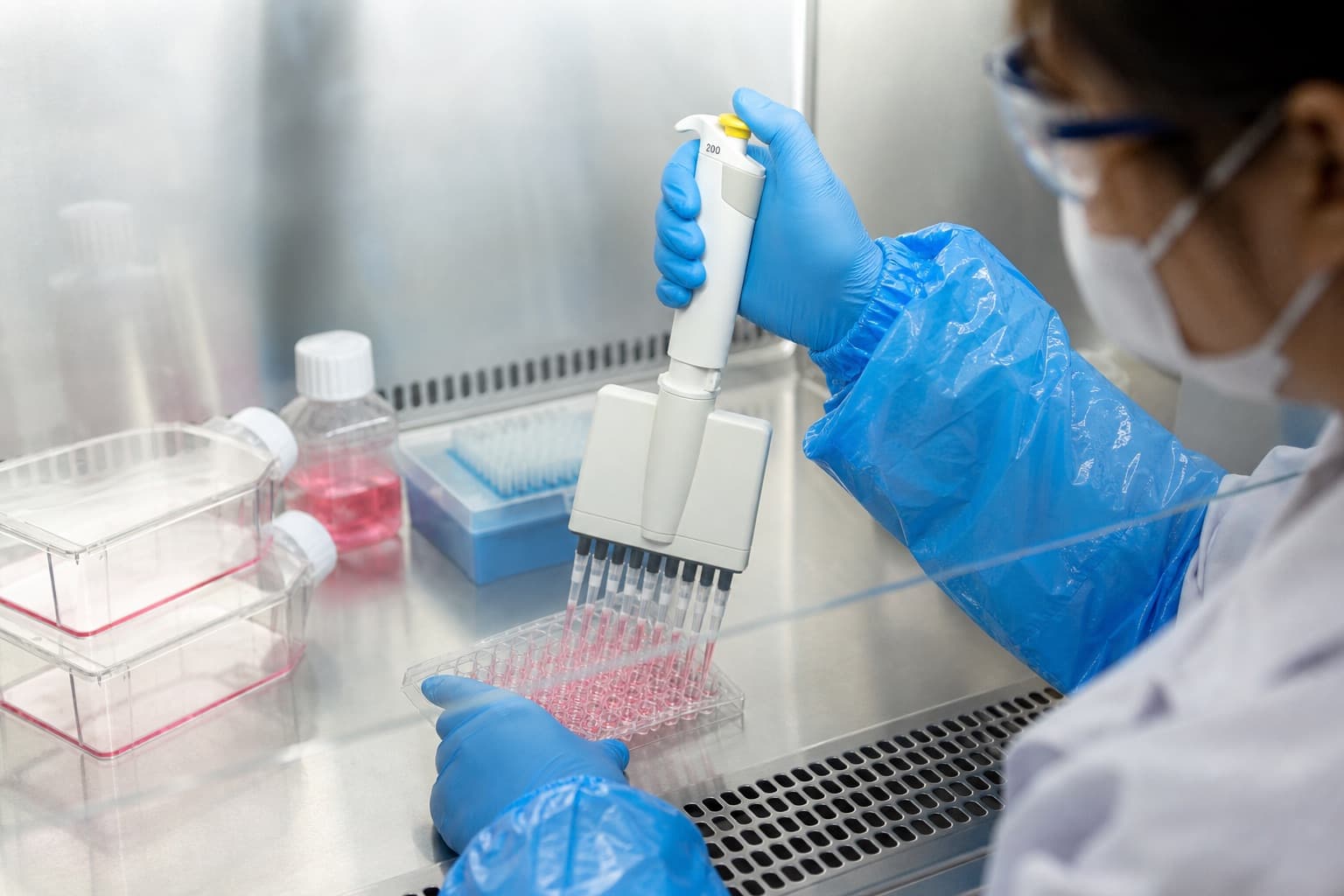A scientist wearing blue sterile sleeves and gloves inside a biosafety cabinet, preparing a sterile bacterial vaccine for infusion. © 2026 ImmunoTherapy Japan. Educational use only